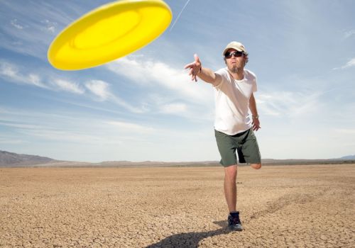 A person in sunglasses and a hat is throwing a yellow frisbee in a dry, cracked desert landscape under a blue sky.