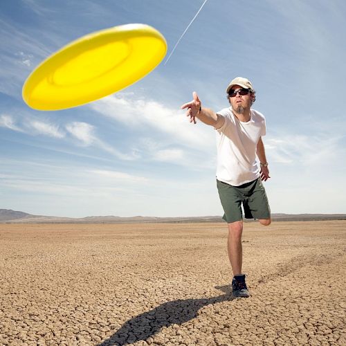 A person in sunglasses and a hat is throwing a yellow frisbee in a dry, cracked desert landscape under a blue sky.