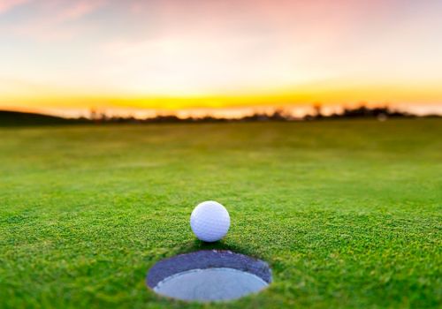 A golf ball is poised at the edge of a hole on a lush green golf course at sunset. The sky displays vibrant colors, creating a serene atmosphere.