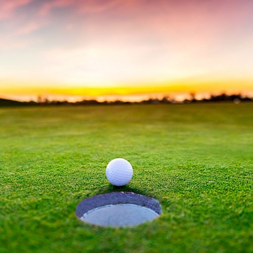 A golf ball is poised at the edge of a hole on a lush green golf course at sunset. The sky displays vibrant colors, creating a serene atmosphere.