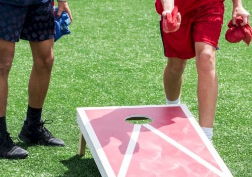 Two people are playing cornhole on a grass field, with one throwing a red bean bag towards the board.