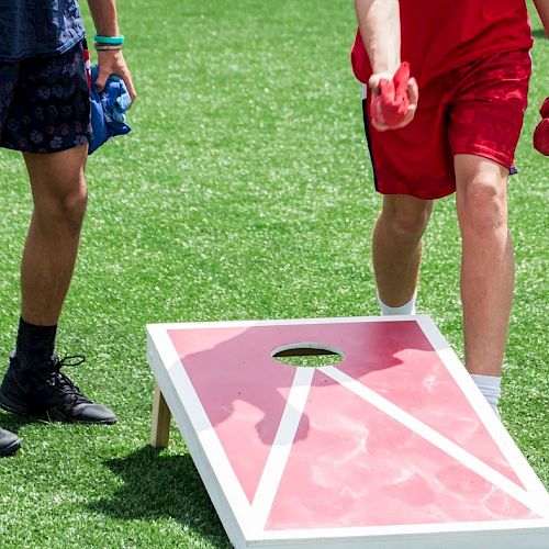 Two people are playing cornhole on a grass field, with one throwing a red bean bag towards the board.