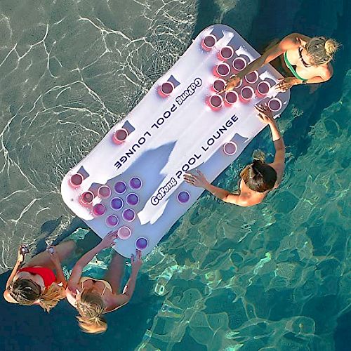 Four people are gathered in a pool around a floating beer pong table with cups arranged on it. They appear to be enjoying a pool party.