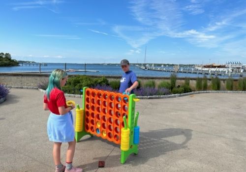 Two people are playing a large outdoor Connect Four game near a waterfront on a sunny day, with boats and a blue sky in the background.