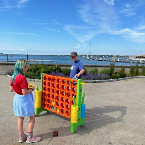 Two people are playing a large outdoor Connect Four game near a waterfront on a sunny day, with boats and a blue sky in the background.