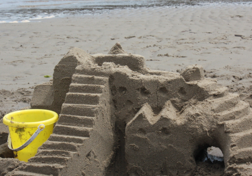 A detailed sandcastle featuring steps, ridges, and an archway sits on the beach near a yellow bucket, with the sea in the background.