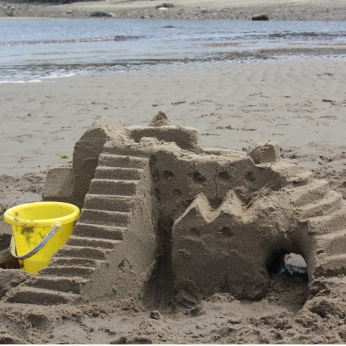 A detailed sandcastle featuring steps, ridges, and an archway sits on the beach near a yellow bucket, with the sea in the background.