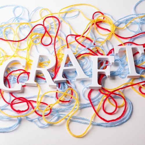 The image shows the word "CRAFT" in white, surrounded by tangled red, yellow, and blue yarn on a white background.