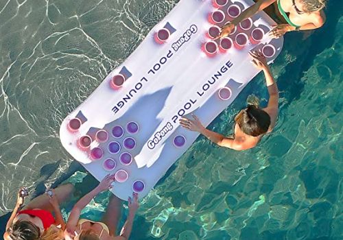 Four people in a pool play beer pong on an inflatable pool lounge with cups arranged on it, enjoying a sunny day.