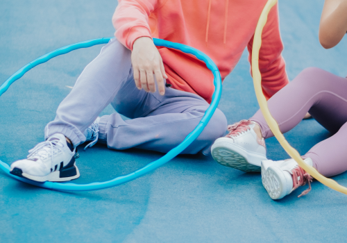 Two people sitting on a blue sports court, each holding a hula hoop, with one person smiling and the other mostly out of the frame.