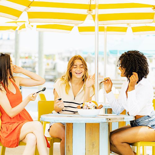 Three people are sitting at an outdoor table under yellow-striped umbrellas, enjoying a shared dessert while laughing and chatting.