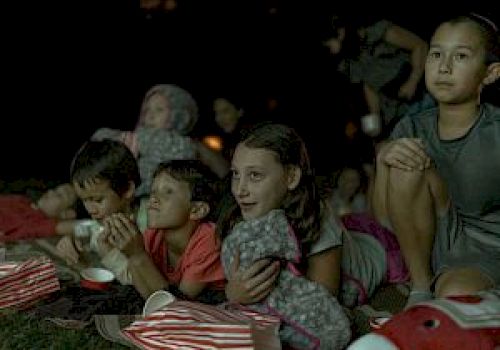 A group of children is lying on the grass with blankets and snacks, watching something intently, possibly an outdoor movie at night.