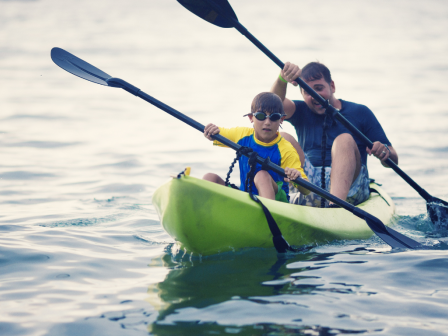 Two people are kayaking together on a body of water, both paddling with oars, with one wearing goggles and a yellow shirt.