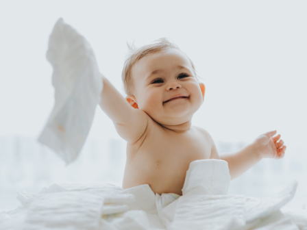 A baby is joyfully playing among white diapers, holding one up with a big smile on their face.