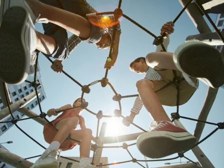 The image shows children climbing on a rope structure at a playground, viewed from below with a bright sky and sunlight in the background.