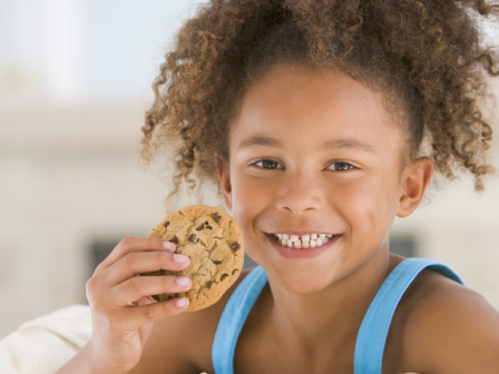 A child with curly hair is smiling and holding a chocolate chip cookie, wearing a blue tank top, appearing to be indoors with a bright background.