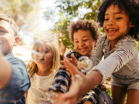 Four children are joyfully playing outdoors, focused on something in front of them, perhaps bubbles. They appear to be in a park or garden.