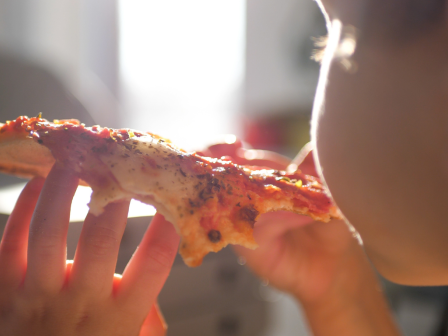 A person is eating a slice of pizza, holding it with both hands. Sunlight is illuminating the scene, creating a warm and appetizing ambiance.