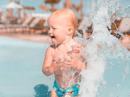 A young child in swimwear plays joyfully near a splash of water at a poolside environment, smiling and enjoying the sunny day.