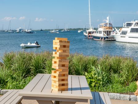 A Jenga tower on a wooden table by a waterfront with boats and yachts in the background, surrounded by greenery and a clear sky.