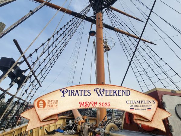 A tall ship&rsquo;s yard, rigging, and a sign reading &ldquo;Pirates Weekend&rdquo; at the ship&rsquo;s bow, with people aboard and a blue sky.