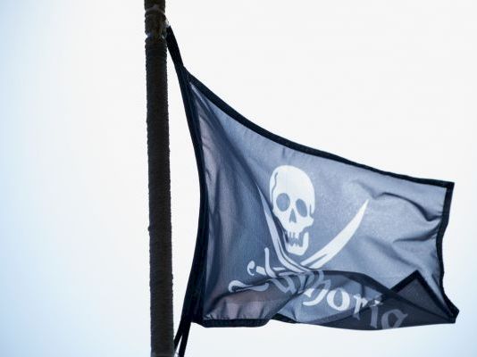 A blue pirate flag with a white skull and crossed swords design, fluttering on a flagpole against the sky.