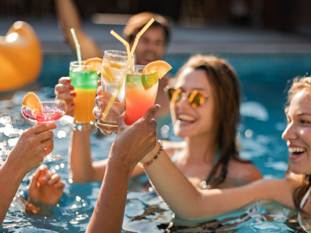 People are in a pool holding colorful cocktails and toasting together, with an inflatable float in the background.