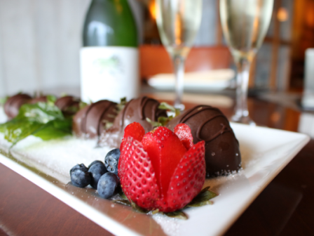 A white plate with chocolate-covered strawberries, blueberries, and a fanned strawberry, accompanied by two champagne glasses and a bottle.