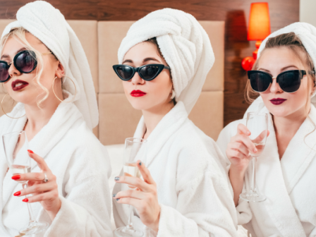 Three women in bathrobes and sunglasses, with towels wrapped around their heads, holding champagne glasses, seated next to a red lamp, seem relaxed.