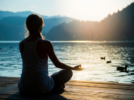 A person is meditating in a seated position by a serene lake, surrounded by mountains and bathed in the glow of the setting sun.