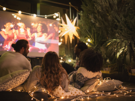 A group of people are watching a movie outdoors at night, surrounded by fairy lights and cozy cushions, with a festive star decoration in the background.