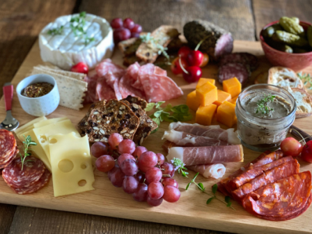 A charcuterie board with various meats, cheeses, grapes, crackers, pickles, nuts, a jar of spread, and mustard in a small dish, on a wooden surface.