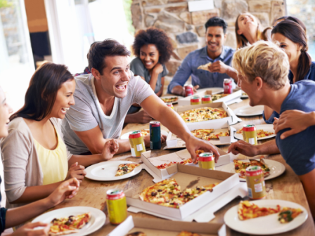 A group of friends is gathered around a table enjoying pizza, laughing, and having a good time together indoors.