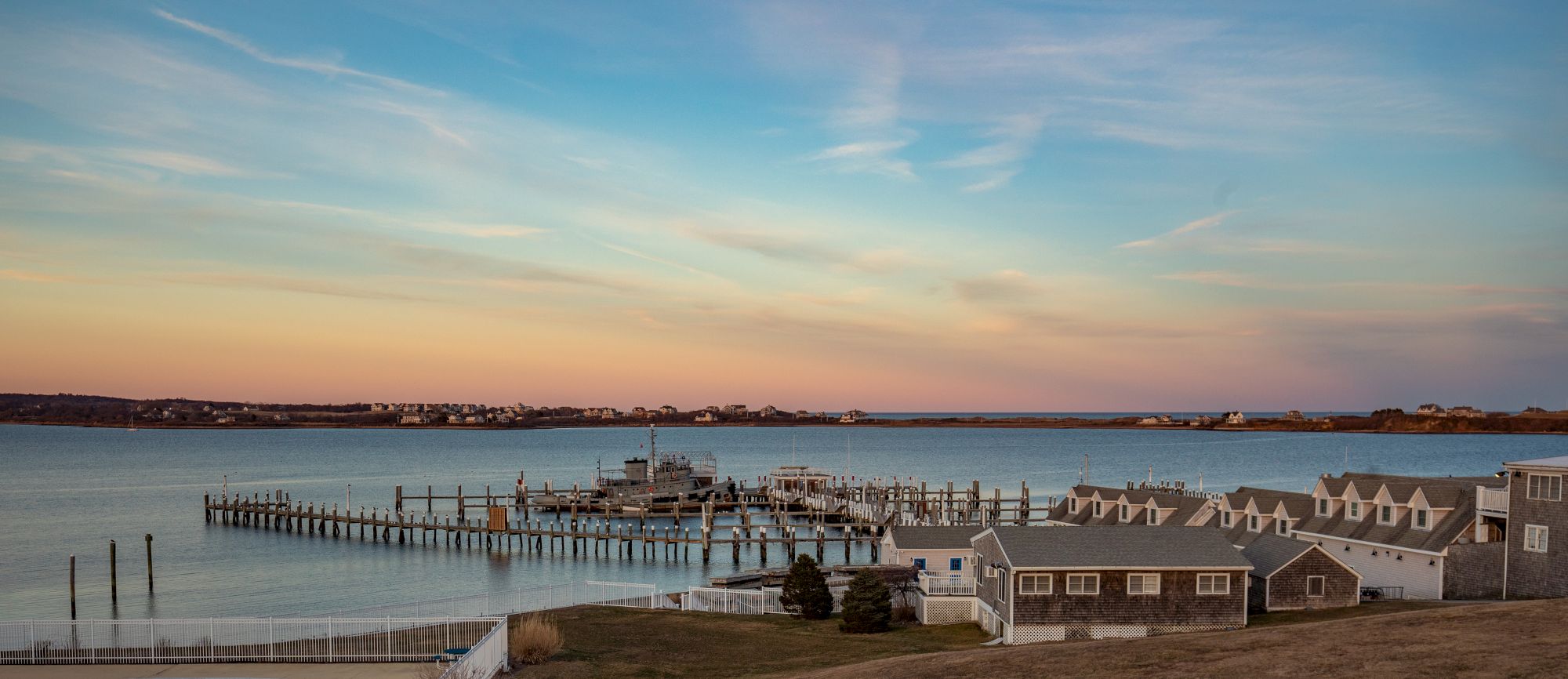 A serene coastal scene with docked boats, houses in the foreground, and a calm sea under a pastel-hued sky during sunset.