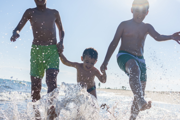 Three children in swim trunks are holding hands, playing, and splashing in the shallow water at the beach under the bright sun.