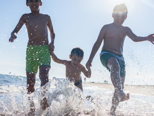 Three children in swim trunks are holding hands, playing, and splashing in the shallow water at the beach under the bright sun.