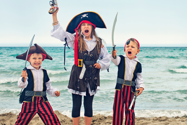 Three children dressed as pirates, holding toy swords and a toy gun, stand on a sandy beach with the ocean in the background.