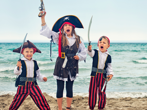 Three children dressed as pirates, holding toy swords and a toy gun, stand on a sandy beach with the ocean in the background.