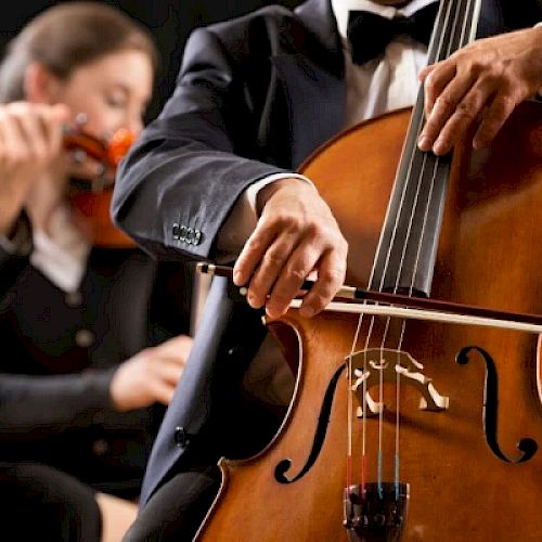 Two musicians are playing string instruments. The focus is on a cellist in the foreground while a violinist is in the background.