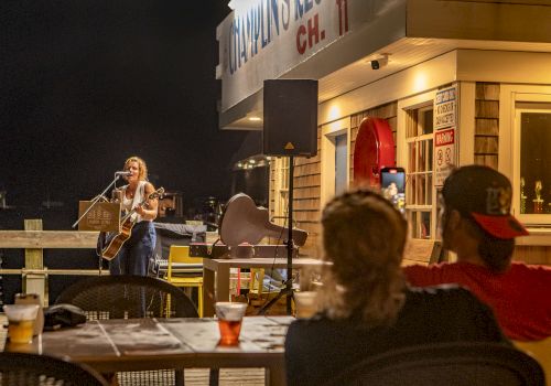 A musician performs on an outdoor stage at night, with a small audience seated at tables enjoying the show, drinks visible on the table.