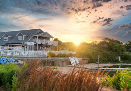 A lakeside house with a deck, golden sunset, and calm waters, framed by reeds and greenery along the shore.