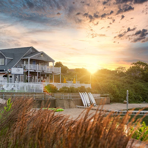 A lakeside house with a deck, golden sunset, and calm waters, framed by reeds and greenery along the shore.
