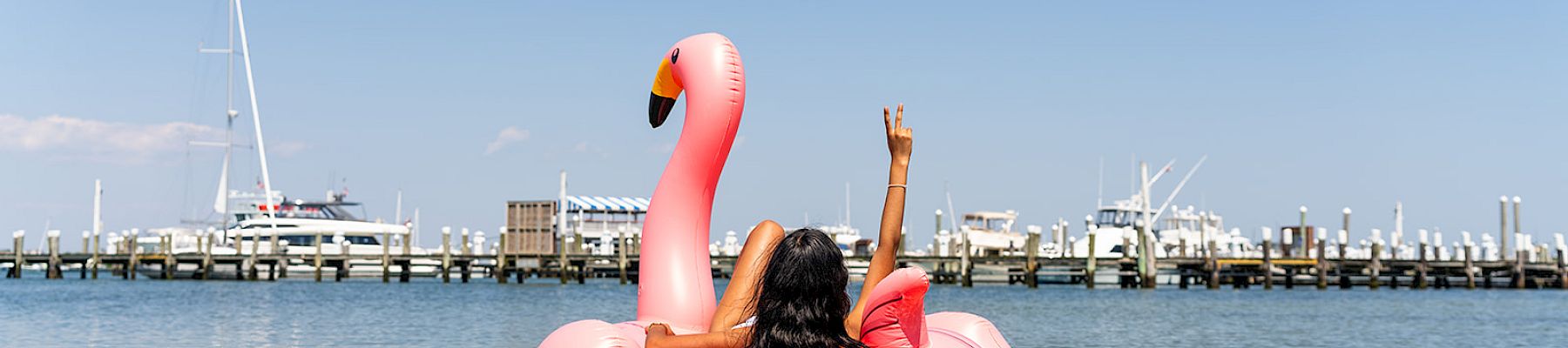 A person relaxes on a pink flamingo float in the water, with a marina and boats in the background, under a clear blue sky.