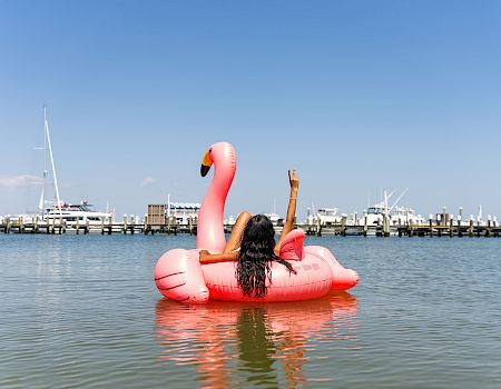 A person relaxes on a pink flamingo float in the water, with a marina and boats in the background, under a clear blue sky.
