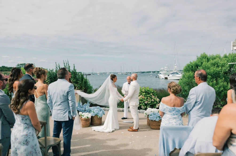 A couple is getting married outside by the water, surrounded by guests in elegant attire, with boats visible in the background.
