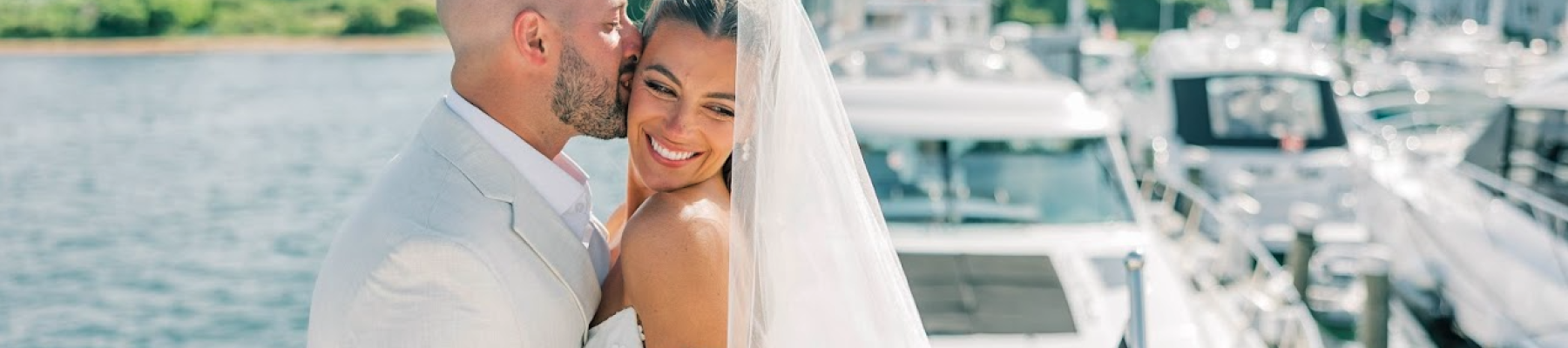 A couple in wedding attire embraces near a dock with yachts in the background, under a clear sky.