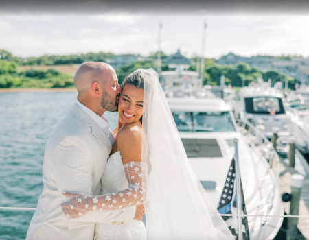 A couple in wedding attire embraces near a dock with yachts in the background, under a clear sky.