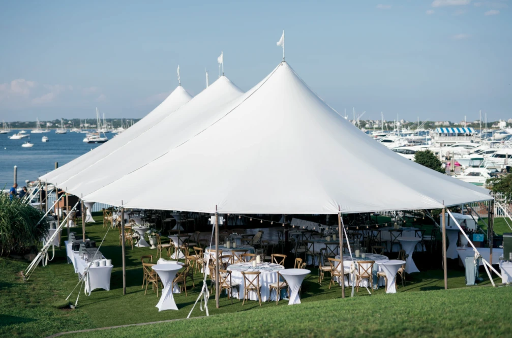 A large white tent is set up on a grassy area near a marina, with tables and chairs underneath, ideal for a waterfront event.