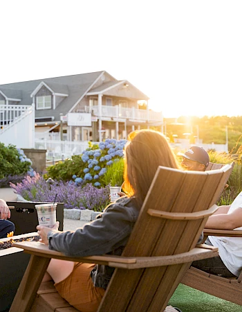 A group of friends lounges in outdoor chairs around a fire pit, chatting and sipping drinks on a sunny patio by the beach houses.