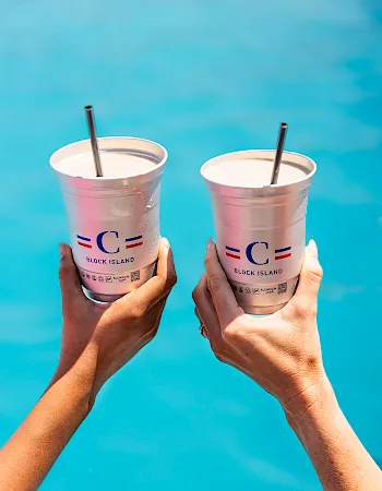 Two hands hold pink iced drinks with straws labeled &ldquo;Block Island&rdquo; against a bright blue pool background.